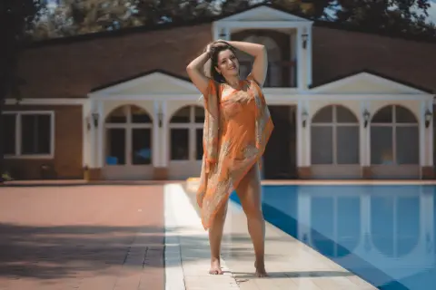 Smiling woman in an orange patterned beach cover-up relaxing by a luxury villa swimming pool.