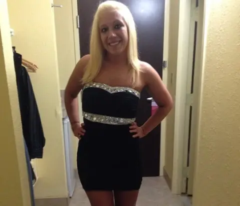 JuliaJay smiles while wearing a short black strapless dress with silver glitter trim, posing with her hands on her hips in a room.