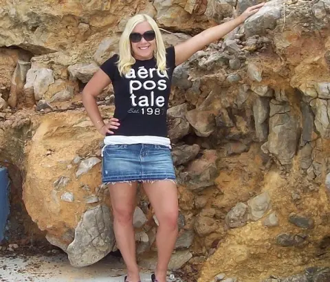 JuliaJay poses outside against a rocky wall, wearing a black tank top, a short denim skirt, and sunglasses.