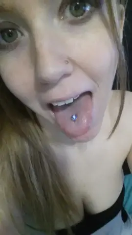 Smiling blonde woman with a nose piercing and tongue stud in a casual indoor setting.