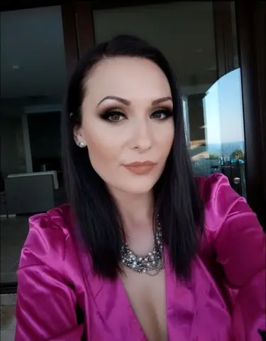 Elegant woman in a pink satin blouse and silver necklace posing with glamorous makeup in a bright setting.