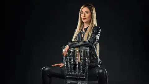 KimDavisLive poses sitting on a clear chair wearing a black leather jacket and black leggings against a dark background.