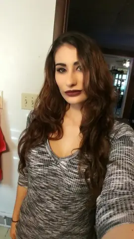 Woman with long wavy hair wearing a grey textured v-neck top in a bright indoor setting.