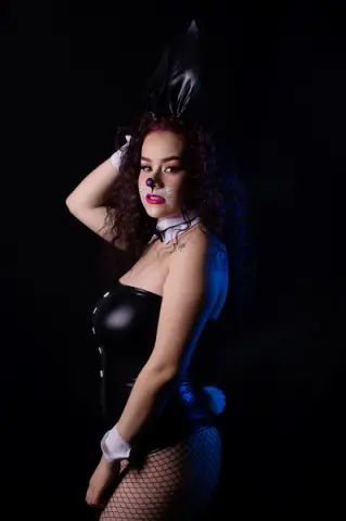 Woman in black bunny costume with ears and fishnet stockings posing against a dark background.