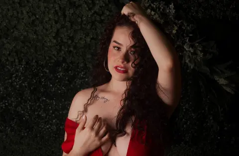 Beautiful woman with curly hair in a red dress posing against a dark green foliage background with a moody vibe.