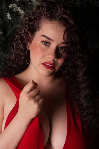 Beautiful woman with curly hair in a red dress posing against a dark floral background with a moody vibe.