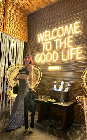 Stylish woman in a black crop top and glittery trousers posing by a neon sign in a cozy, warm lounge setting.