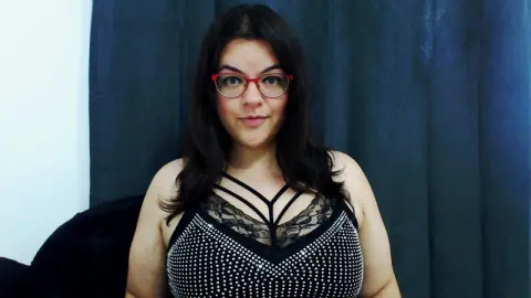 Smiling woman with dark hair and red glasses wearing a black sequined camisole against a blue curtain background.