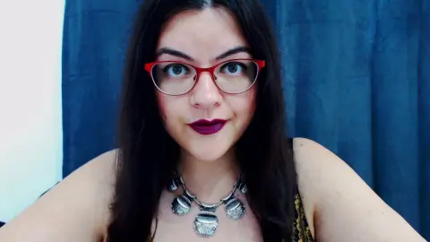 Young woman with red glasses and silver statement necklace smiling against a blue curtain background.