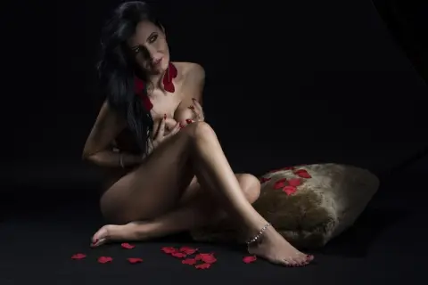 MissyJolie poses naked against a black background, leaning back against a brown furry pillow with red rose petals scattered on the floor.