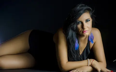 MissyJolie poses lying down against a black background, wearing a black top and blue feather earrings.