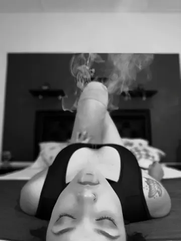 Relaxed woman in black sports bra lying on bed amidst dreamy swirling smoke in a moody black and white setting.