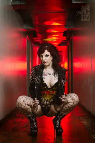 Gothic woman in black leather jacket and fishnets squatting in a moody, red-lit industrial hallway.