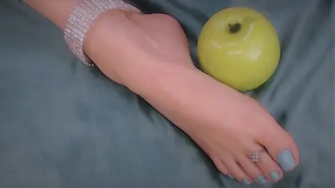 Close-up of a woman's bare foot with blue nail polish resting next to a green apple on a soft teal fabric.