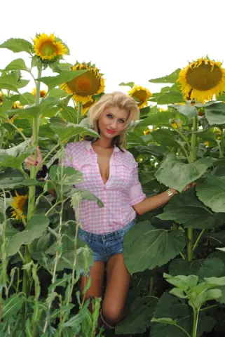 Blonde woman in a pink gingham shirt and denim shorts smiling in a sunny sunflower field.