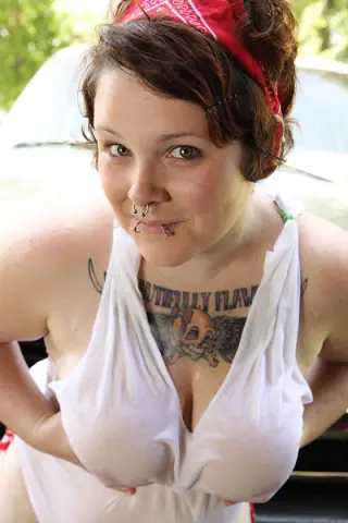 Smiling woman with nose ring and tattoos wearing a white tank top in a bright outdoor setting.
