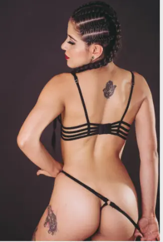 Woman with braided hair and back tattoos wearing black minimalist lingerie against a dark studio background.