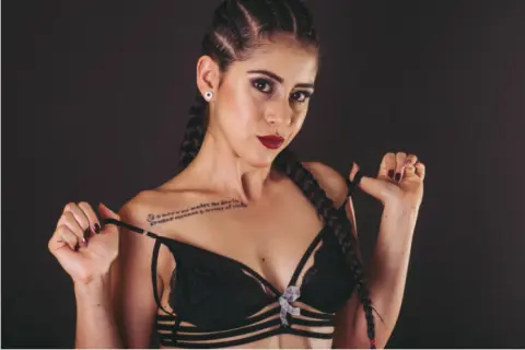 Young woman with braided hair in black lace lingerie posing confidently against a dark studio background.