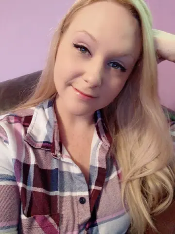 Smiling blonde woman in a plaid flannel shirt relaxing against a pink background.