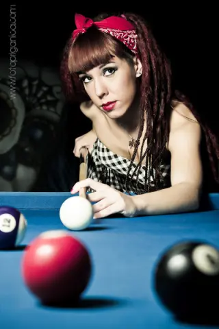 Sofi_Mora with red hair and a headband leans over a blue pool table, holding a cue stick near the white ball.