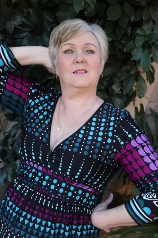 Smiling woman in a patterned teal and black wrap dress posing against a lush green leafy background.
