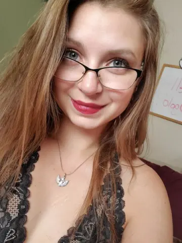 Smiling young woman with glasses and a black lace top in a cozy indoor setting.