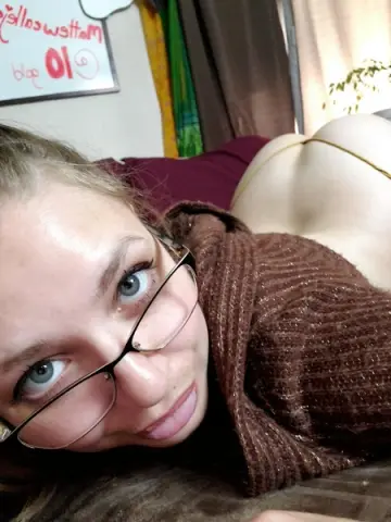 Young woman with glasses in a brown sweater relaxing on a bed in a cozy indoor setting.