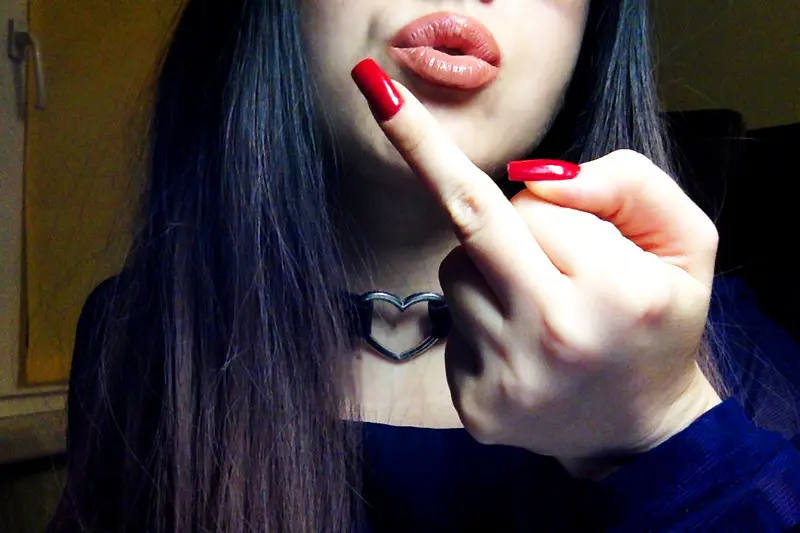 Rebellious young woman with long dark hair and red nails making a rude gesture in a moody indoor setting.