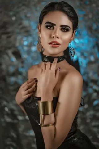 Glamorous woman in a black strapless dress and gold cuff jewelry posing against a shimmering blue background.