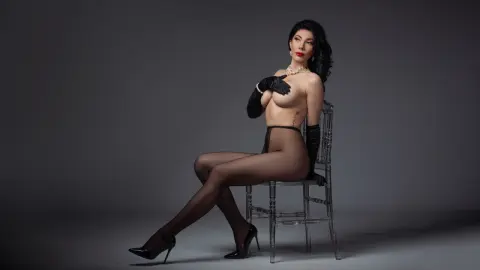 Elegant woman in black gloves and sheer tights posing on a clear chair against a grey studio background.