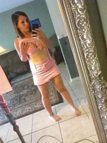 Woman in a pink crop top and mini skirt taking a mirror selfie in a modern living room.