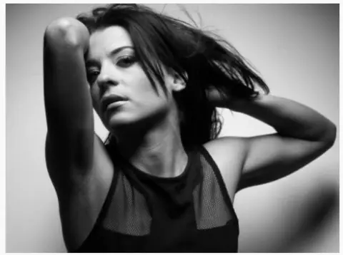 Moody black and white portrait of a woman in a black mesh tank top posing against a plain background.