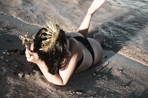 Mermaid in black bikini wearing a gold crown lying on a sandy beach by the ocean waves.