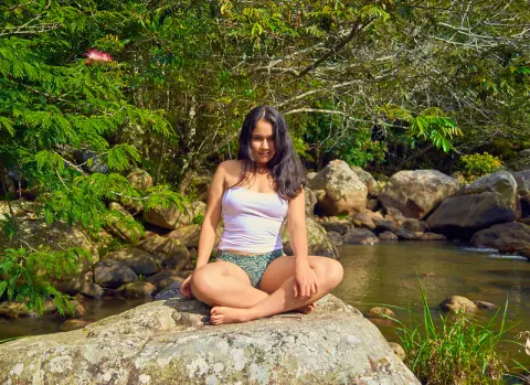 Young woman in a white tank top and patterned bikini sitting on rocks by a peaceful forest river.