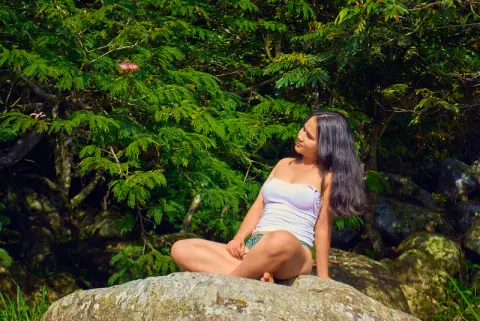 Young woman in a white tank top sitting on rocks amidst lush green forest foliage in a peaceful outdoor setting.